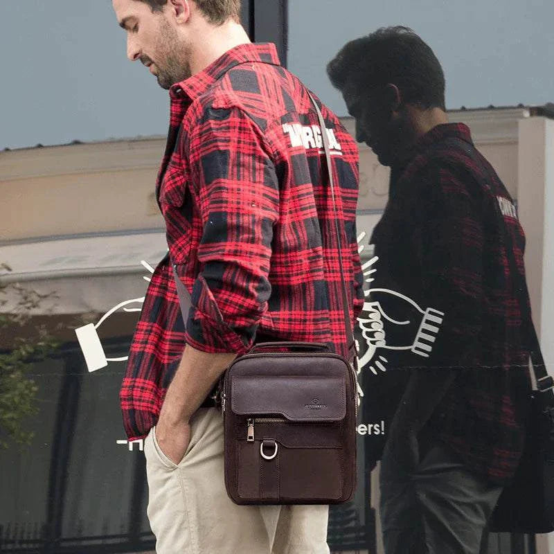 Man carrying a vintage leather messenger bag with a distressed finish, wearing a red plaid shirt and beige pants.